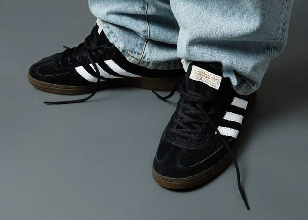 Black Adidas sneakers with white stripes worn with light blue jeans on a gray background